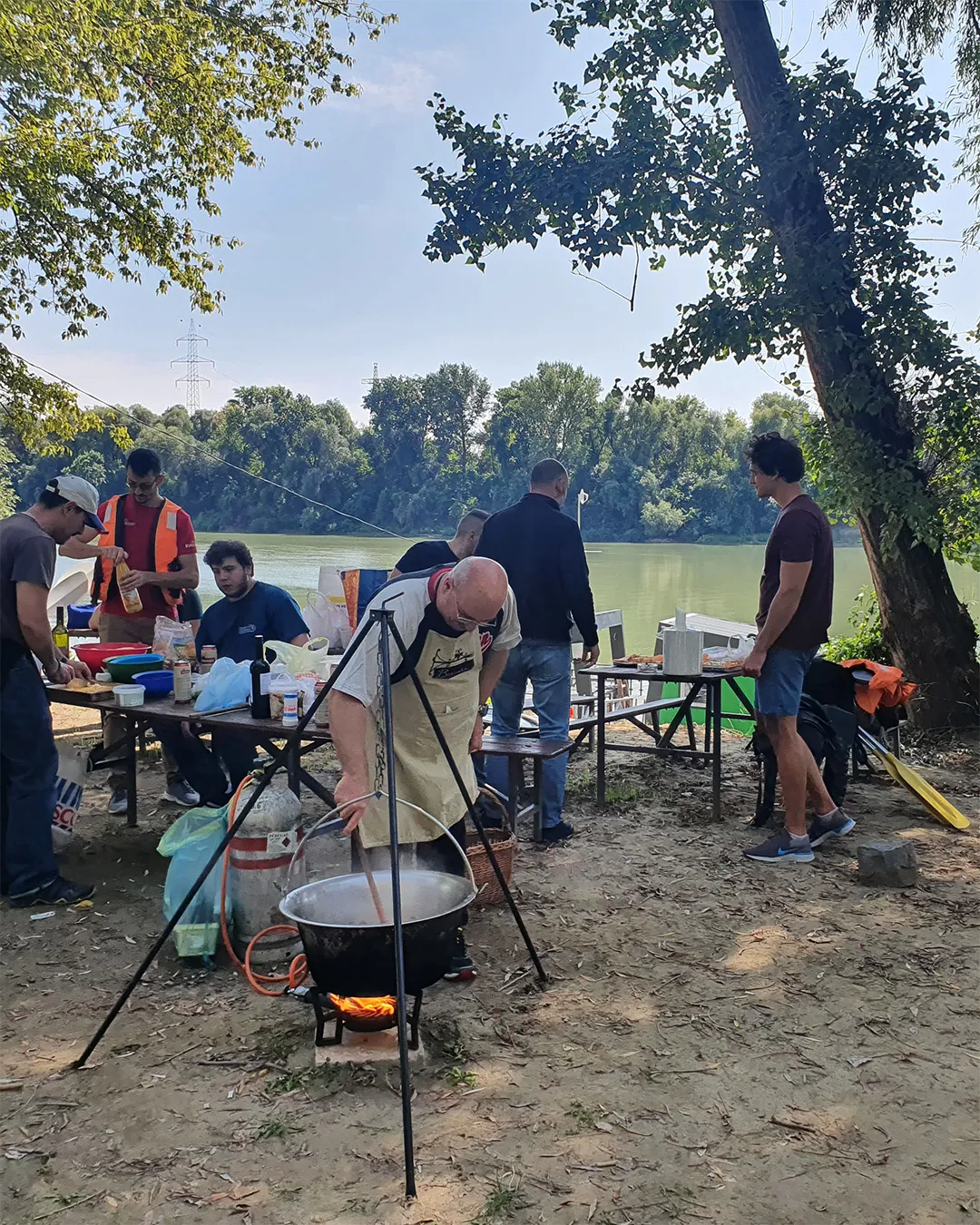 A csapat a Tisza partján: bográcsban fő az ebéd, a háttérben a folyó és a kikötött csónakok evezői.